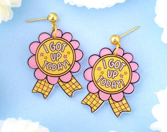 May include: A pair of pink and yellow earrings shaped like a ribbon with a flower and the text "I got up today!" in the center.