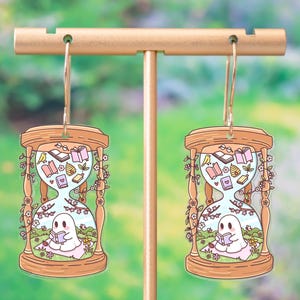 May include: A pair of whimsical hourglass earrings. Each earring features a wooden-framed hourglass design with a ghost reading a book in a garden scene. The hourglass is filled with books, flowers, and a bird. The earrings hang from gold-colored hooks.