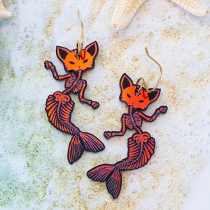 May include: A pair of orange and black skeleton cat mermaid earrings. The earrings are made of acrylic and have a shiny finish.