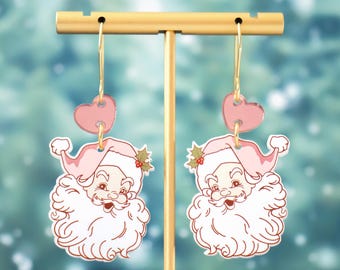 May include: A pair of pink and white Santa Claus earrings with a pink heart charm. The earrings are hanging from a gold stand.