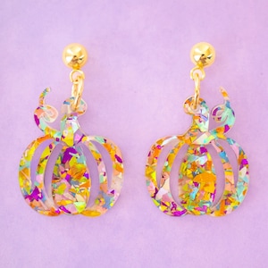 May include: A pair of gold pumpkin earrings with iridescent glitter embedded in the acrylic. The earrings are shaped like pumpkins with a stem and a swirl design.