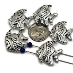 May include: Silver-colored fish-shaped pins with detailed scales and fins. A dime is included for size reference. The pins have a blue bead at the base of the pin. The fish pins are arranged in a circular pattern.