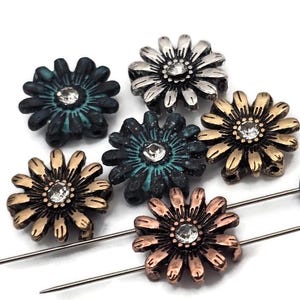May include: Six decorative flower-shaped pins in various metallic finishes: silver, bronze, and copper, with a central clear rhinestone. The pins are arranged on a white background, showcasing their intricate petal details and sparkling centers.