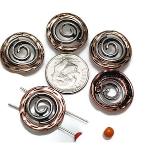 May include: Six metal spiral-design beads in copper and silver tones, with a dime for size comparison. The beads have a textured outer ring and a swirling silver center. A small orange bead and two pins are also visible.