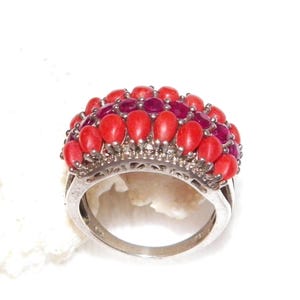 May include: A silver ring featuring a dome-shaped cluster of oval-cut red coral cabochons and a central row of deep pink faceted stones. The metal band has a decorative filigree gallery setting that supports the multi-layered gemstone arrangement.