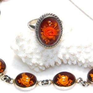 May include: A silver ring and bracelet set featuring oval-shaped amber-colored resin stones with orange rose designs. The ring has a braided silver band. The bracelet has multiple oval stones connected by silver links. The jewelry is displayed on a white surface.
