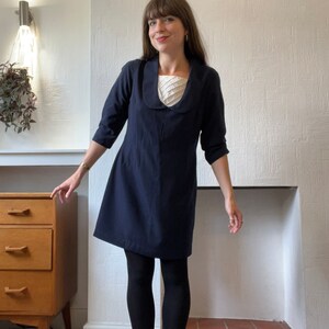 May include: A woman wearing a navy blue dress with a white collar and white lace trim. She is standing in front of a white wall with a wooden dresser behind her.