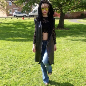 Könnte beinhalten: Eine Frau trägt eine lange schwarze Kunstfellweste mit einem schwarzen Spitzenoberteil und blauen Jeans. Sie trägt eine gelbe Sonnenbrille und hat ihre Haare nach hinten gebunden.