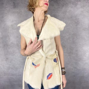 Puede incluir: Chaleco de piel sintética color crema con cuello ancho y cierre de lazo. El chaleco presenta parches con diseños de bandera americana y labios. La modelo lleva vaqueros azules y un collar de cuentas. Una prenda de abrigo sin mangas con estilo.