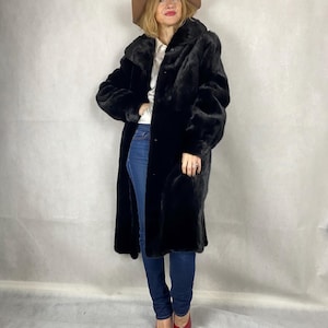 May include: A long black fur coat with a hood and a button fastening. The coat is being worn by a person wearing a white shirt, blue jeans, and red shoes with a jeweled embellishment.