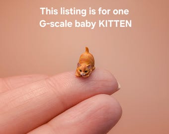 May include: A miniature orange tabby kitten figurine in a crouching position. The text above the kitten reads: This listing is for one G-scale baby KITTEN. The kitten has dark orange stripes, a light-colored muzzle, and small pointed ears.