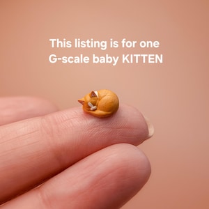 May include: A miniature orange G-scale baby kitten figurine in a curled sleeping position. The text above the kitten reads: This listing is for one G-scale baby KITTEN. The kitten features small white markings on its face and ears.