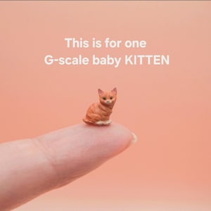 May include: A miniature G-scale orange tabby kitten figurine with pointed ears and a seated posture. The text above the kitten reads: This is for one G-scale baby KITTEN. The figurine is rendered in shades of orange and white with detailed facial features.