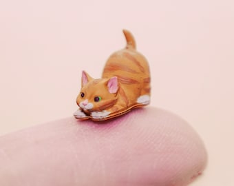 Pode incluir: Uma pequena figura de gato laranja e branco em uma pose brincalhona. O gato está deitado de bruços com a cabeça virada para o lado e as patas esticadas.