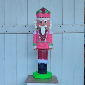May include: A decorative, colorful nutcracker figurine standing on a white pedestal. The nutcracker is dressed in a pink uniform with a red and green hat. It has a white mustache and beard, and is standing on a green base.