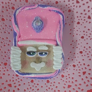 May include: A pink and purple glittery gingerbread cookie with a white icing crown and a sparkly blue gem. The cookie has a brown face with white icing details and green and blue eyes.