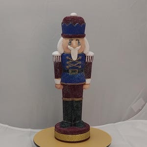 May include: A decorative nutcracker figurine in a traditional soldier design. The figurine features a blue and red uniform with white hair and a long, white mustache. It stands on a gold and black base.