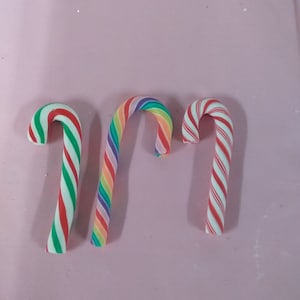 May include: Three candy canes, one red and white, one rainbow, and one red and white. The candy canes are on a pink background.