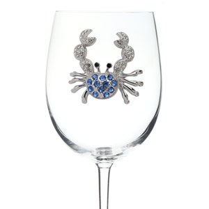 May include: A clear glass wine glass with a silver crab charm. The crab is decorated with blue rhinestones and has a silver charm attached to the bottom of the glass that says "The Glimmering Jewels".