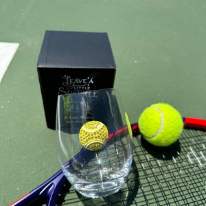 May include: A clear glass with a yellow tennis ball design on it. The glass is sitting on a tennis racket with a tennis ball next to it. The glass is part of a set that includes a box with the text "Leave a Little Sparkle" on it.