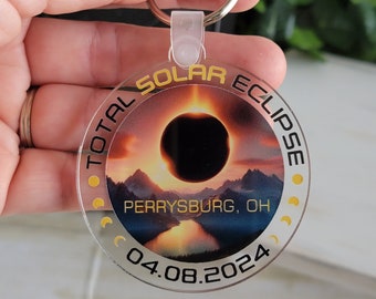 May include: A round, clear acrylic keychain with a gold border and the text "TOTAL SOLAR ECLIPSE" and "PERRYSBURG, OH 04.08.2024". The keychain features a photo of a total solar eclipse with mountains and a lake in the foreground.