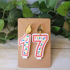 A pair of earrings designed to resemble birthday candles in the shape of number 17, displayed on a brown card The earrings feature white base with red outline and colorful sprinkle-like pattern Above each number is a gold flame-shaped attachment.