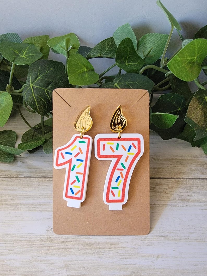 A pair of earrings designed to resemble birthday candles in the shape of number 17, displayed on a brown card The earrings feature white base with red outline and colorful sprinkle-like pattern Above each number is a gold flame-shaped attachment.