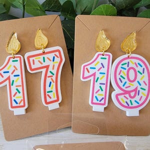May include: A pair of birthday candle earrings with gold tops and colorful sprinkles. The earrings are shaped like the numbers 17 and 19.