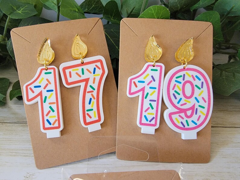 May include: A pair of birthday candle earrings with gold tops and colorful sprinkles. The earrings are shaped like the numbers 17 and 19.