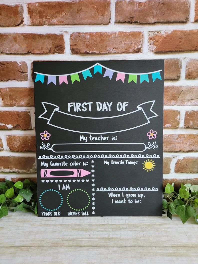 May include: Black chalkboard with a white banner that says "FIRST DAY OF". The chalkboard has spaces to fill in information about the first day of school, including the student's teacher, favorite color, favorite things, age, height, and what they want to be when they grow up.