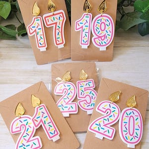 May include: Four pairs of birthday candle earrings. Each pair features a different number, 17, 19, 21, and 20. The candles are white with pink frosting and colorful sprinkles. The earrings have gold tops.