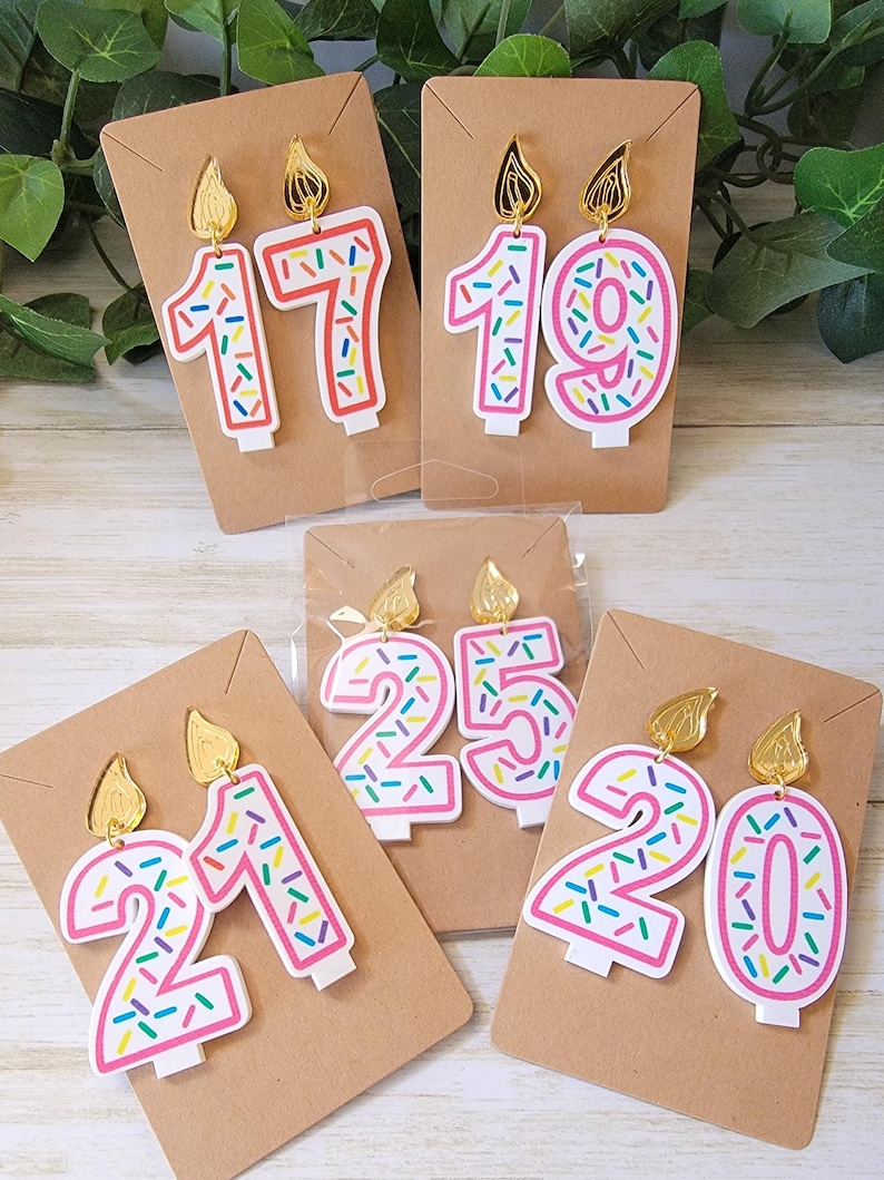 May include: Four pairs of birthday candle earrings. Each pair features a different number, 17, 19, 21, and 20. The candles are white with pink frosting and colorful sprinkles. The earrings have gold tops.