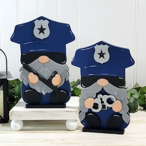 May include: Two wooden gnome figurines dressed as police officers. The larger gnome is holding a nightstick and the smaller gnome is holding handcuffs. Both gnomes are wearing blue hats with a silver star and sunglasses.