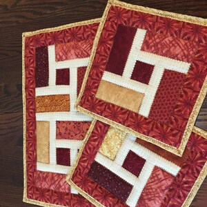 May include: Three handmade quilted placemats with a geometric design. The placemats feature a patchwork of red, burgundy, and tan fabrics, with white accents. Each placemat has a decorative border and is placed on a dark wood surface.