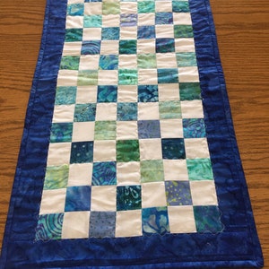 May include: A rectangular quilted table runner with a checkerboard pattern of blue, green, and white squares. The runner has a solid blue border with a scalloped edge, and is displayed on a wooden surface.