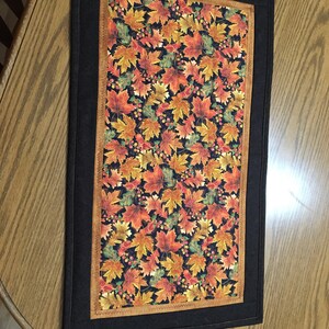 Peut inclure: Une nappe rectangulaire avec un motif de feuilles d'automne orange, rouge et jaune sur fond sombre. Le motif de feuilles est encadré par une bordure marron et une bordure extérieure noire. La nappe est probablement en tissu.
