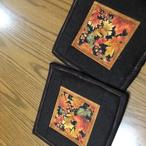 May include: Two square black quilted pot holders featuring a central square of autumn leaves in orange, yellow, and green. The leaves are framed by a brown border and stitched with a decorative stitch.