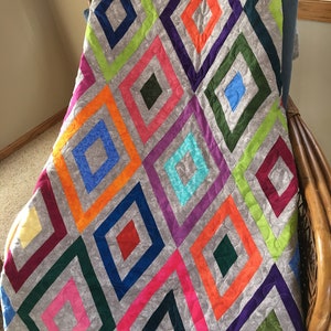 May include: A colorful quilt with a geometric diamond pattern. The quilt features a variety of vibrant colors including pink, orange, blue, green, and purple, set against a gray background. The quilt is draped over a wicker chair.