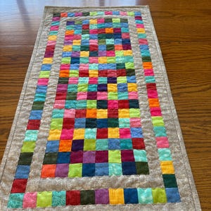 May include: A colorful patchwork table runner with a gray border. The runner is made of squares of different colors, including red, orange, yellow, green, blue, purple, and pink.
