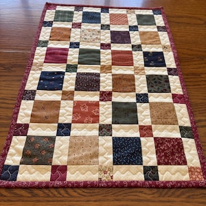 May include: A small, rectangular patchwork quilt with a grid pattern. The quilt features squares of various colors and patterns, including floral and geometric designs. The border is a solid burgundy colour, and the quilt is placed on a wooden surface.