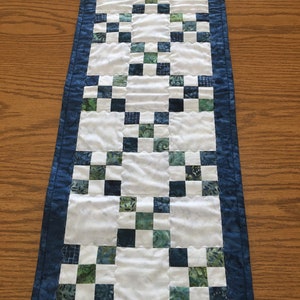 May include: A rectangular quilted table runner featuring a patchwork design. The runner has a white center with alternating squares of blue and green fabric. A dark blue border frames the design, adding a contrasting edge to the handmade textile.