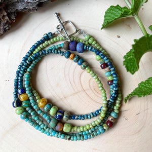 May include: A multi-strand necklace made of small, colorful glass beads. The necklace features shades of blue, green, and yellow. The beads are strung on a silver chain with a clasp.