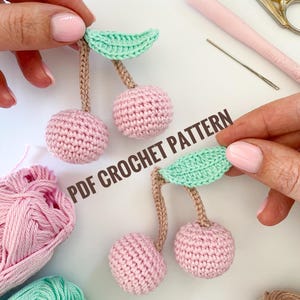 May include: Two crocheted cherries with green leaves. The cherries are pink and the leaves are light green. The cherries are attached to brown stems. The image also shows balls of pink and green yarn.