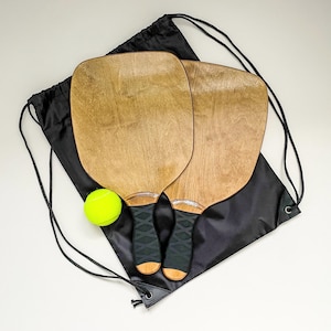 Op de afbeelding: Twee houten pickleball rackets met zwarte handgrepen en een gele pickleball liggen op een zwarte trekkoordtas. De rackets hebben een lichtbruine houtnerf afwerking. De tas is gesloten met een zwart trekkoord.