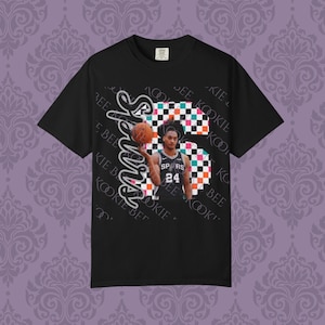 Pode incluir: Camiseta preta com um jogador de basquete e o número 24. A camisa tem um padrão xadrez e a palavra "Spurs". O texto "Kookie Bee" é impresso na camisa em um padrão repetitivo.