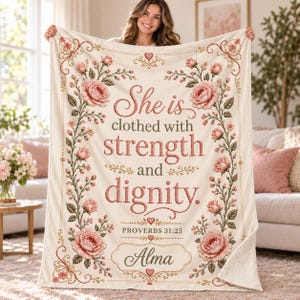 Peut inclure: Une couverture en polaire crème ornée d'une bordure florale de roses roses et de feuilles vertes. Le texte indique : "She is clothed with strength and dignity. PROVERBS 31:25" avec le prénom "Alma" centré en bas dans un cadre décoratif.