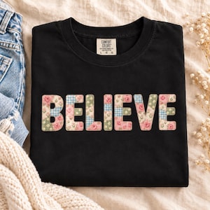 Può includere: Una t-shirt nera a maniche corte con la parola BELIEVE stampata in grandi lettere maiuscole sul petto. Le lettere presentano un design patchwork di rose rosa, foglie verdi e motivi a quadretti blu e bianchi su sfondo color crema.