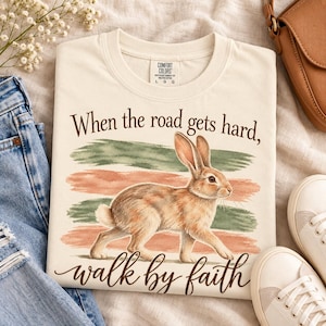 Peut inclure: Un t-shirt à manches courtes de couleur crème avec une illustration de lapin brun centrée sur des coups de pinceau horizontaux verts et pêche. Le texte indique When the road gets hard, walk by faith. Étiquette Comfort Colors au col.