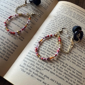 May include: A pair of black, geometric earrings with gold-colored hooks and chains, connected to a beaded bracelet with red, pink, and gold beads. The jewelry rests on an open book.