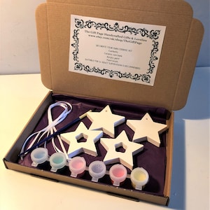 May include: A craft kit for decorating ceramic stars. The kit includes five white ceramic stars, a paintbrush, and six small bottles of acrylic paint in various colors. The kit is packaged in a brown cardboard box with a white label that reads "The Gift Page Handcrafted Gifts & Lovelies" and "DECORATE YOUR OWN CERAMIC KIT".